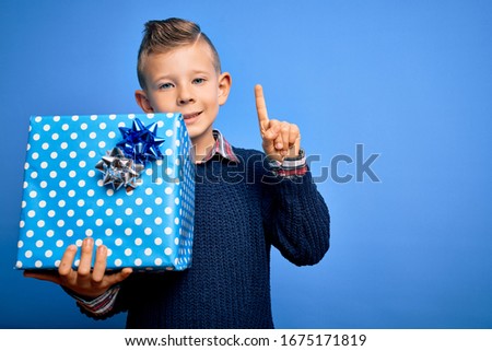 Young little caucasian kid holding surprise box as birthday or Christmas present surprised with an idea or question pointing finger with happy face, number one