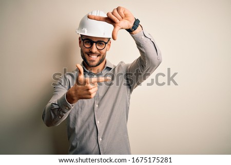 Young architect man wearing builder safety helmet over isolated background smiling making frame with hands and fingers with happy face. Creativity and photography concept.