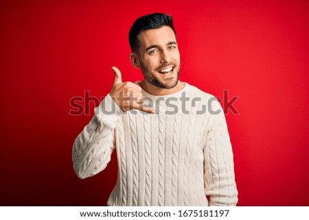 Young handsome man wearing casual white sweater standing over isolated red background smiling doing phone gesture with hand and fingers like talking on the telephone. Communicating concepts.