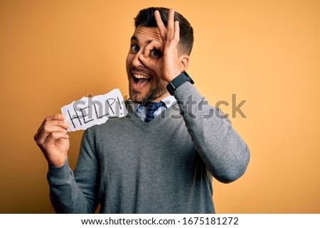 Young overworked business man asking for help holding paper over yellow background with happy face smiling doing ok sign with hand on eye looking through fingers