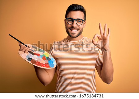 Young handsome artist man with beard painting using bursh and palette with colors doing ok sign with fingers, excellent symbol