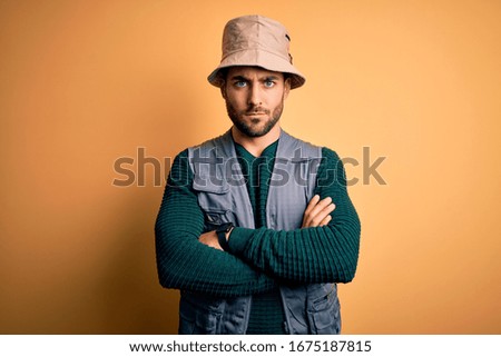 Handsome tourist man with beard on vacation wearing explorer hat over yellow background skeptic and nervous, disapproving expression on face with crossed arms. Negative person.