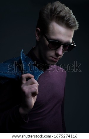 Closeup portrait of a young guy, looking at the camera, holding a denim jacket on his shoulder. Denim style. on a gray background.