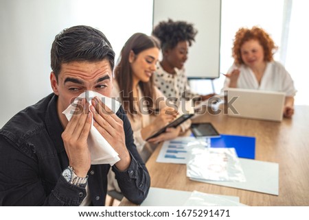 Young man blowing his nose while working. Young sick businessman blowing his nose while working in the office. There are people in the background. Businessman blowing his nose with a tissue at work