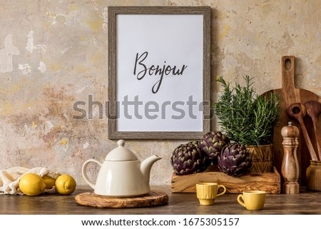 Stylish interior of kitchen space with wooden table, brown mock up photo frame, lemons, vegetables, tea pot, cups and kitchen accessories in wabi sabi concept of home decor. 
