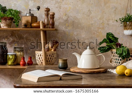 Stylish composition of kitchen interior with family table, vegetables, tea pot, dessert, shelf with food supplies, plants and kitchen accessories in wabi sabi concept of home decor. 