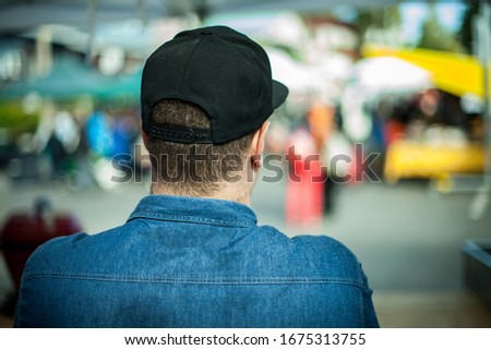 A large build man is seen from behind in selective focus, wearing blue denim shirt and black baseball cap at a street market with blurry stalls in background.