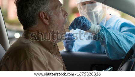 A medical technician in full protective gear collects a sample from a mature man sitting inside his car as part of the operations of a coronavirus mobile testing unit.
