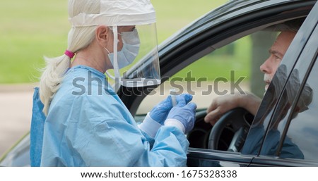 Dressed in full protective gear a healthcare worker collects a sample from a mature man sitting inside his car as part of the operations of a coronavirus mobile testing unit. Royalty-Free Stock Photo #1675328338