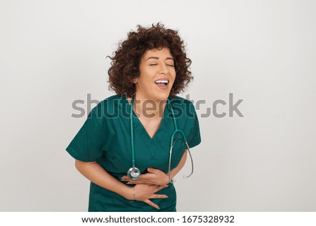 Joyful Caucasian doctor woman wearing medical uniform keeps hands crossed, laughs at good joke, wears casual clothes and round spectacles, standing indoors.