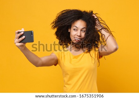 Stunning young african american woman girl in casual t-shirt posing isolated on yellow orange background in studio. People lifestyle concept. Mock up copy space. Doing selfie shot on mobile phone