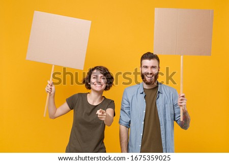 Funny protesting two people guy girl hold protest signs broadsheet blank placard on stick pointing finger on camera isolated on yellow background. Protests strikes pickets concept. Youth against city