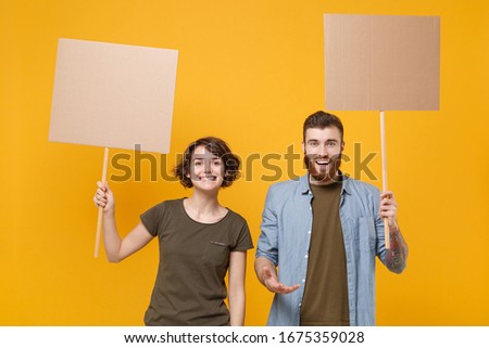 Smiling protesting young two people guy girl hold protest signs broadsheet blank placard on stick isolated on yellow background studio portrait. Protests strikes pickets concept. Youth against city