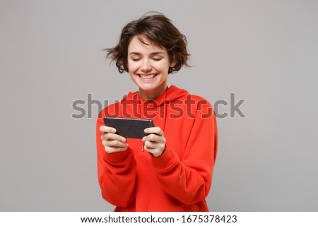 Cheerful young brunette woman girl in casual red hoodie posing isolated on grey background studio portrait. People sincere emotions lifestyle concept. Mock up copy space. Play game with mobile phone