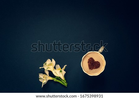 Minimalistic flat lay. Several buds of flowers and coffee with cream and cinnamon in the form of a heart in a mug on a dark blue background. White with yellow petals. Vignetting. Copy space on top.