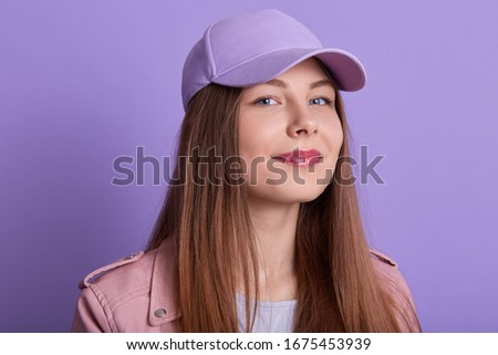 Picture of beautiful young pleasant blonde girl, lady wearing cap and jacket, fashionable girl looking directly at camera, posing isolated over lilac studio background, looks happy and satisfied.