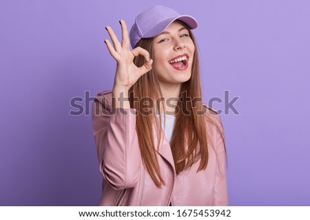 Horizontal portrait of magnetic pretty attractive female standing isolated over lilac background in studio, closing one eye, wearing cap and leather jacket, opening mouth widely, showing sign ok.