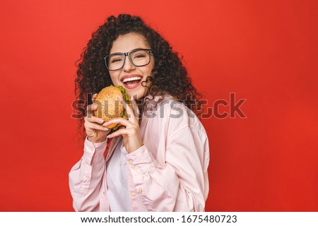 Portrait of young curly beautiful hungry woman eating burger. Isolated portrait of student with fast food over red background. Diet concept. 