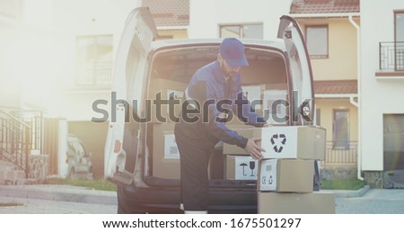 Caucasian male courier in the blue costume and a cap taking out mail carton boxes from the white van on the sunny day in the street. Outdoor.