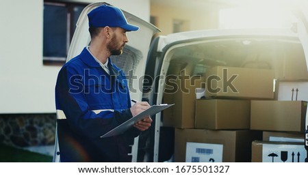 Caucasian handsome mailman in the blue uniform and cap standing at the white van with carton boxes and filling in documents on the clipboard. Outdoors. Royalty-Free Stock Photo #1675501327