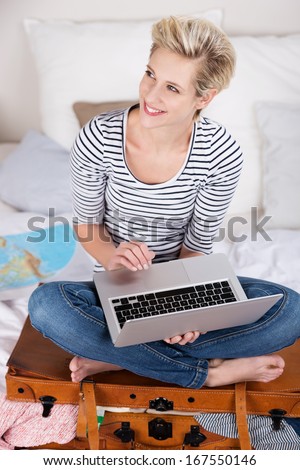 Excited woman daydreaming of her annual vacation sitting on her suitcase on a bed with her laptop in her hands smiling as she looks up off to the left of the frame