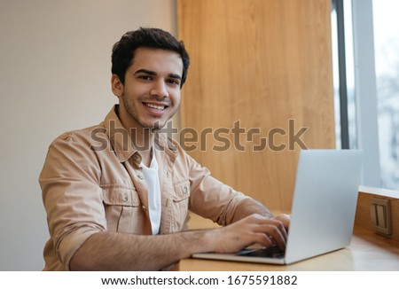 Portrait of happy successful manager using laptop computer, working online project. Handsome Indian man typing on keyboard, searching on website, looking at camera and smiling