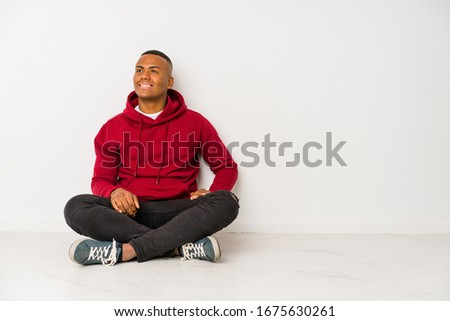 Young latin man sitting on the floor isolated looks aside smiling, cheerful and pleasant.