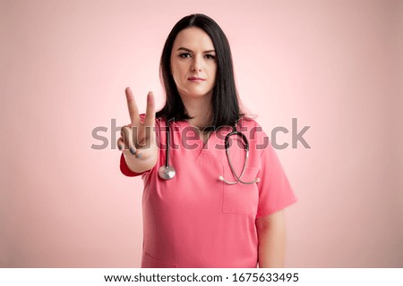 Portrait of beautiful woman doctor with stethoscope, wearing pink scrubs, showing victory sign posing on a pink isolated backround.
