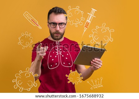 Young bearded man in trendy hipster eyeglasses holding tablet and pointing directly at the camera with his index finger with serious face expression warning about coronavirus danger. 