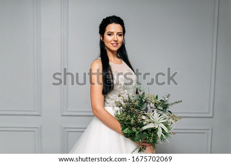 Portrait of a charming young woman in a wedding dress, she holds a bouquet in her hands and looks at the camera. Studio photo with gray background