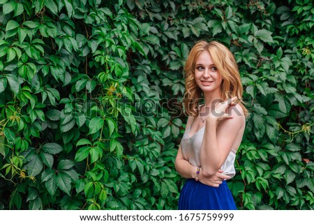 Portrait of a young beautiful woman with make up and curly blond hair, standing next to the wall of wild grape leaves. Copy space.