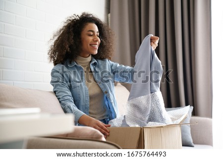 Young curly hair satisfied happy african girl woman lady shopaholic customer sit on sofa unpack parcel delivery box look at clothes take out grey pullover sweater, online shopping shipment concept. Royalty-Free Stock Photo #1675764493