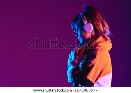Happy igen teen hipster fashion girl model wear stylish glasses headphones enjoy listen new cool trance music mix stand at purple studio background in trendy 80s 90s club party light, profile view Royalty-Free Stock Photo #1675809577