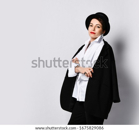 Young woman in stylish boater hat and white shirt blouse wearing black jacket over her shoulders is posing with her arms crossed over white background with text copy space