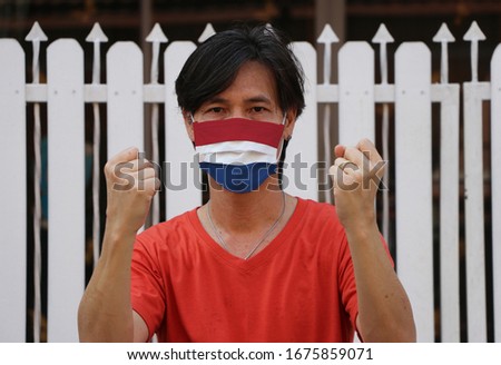 Netherlands flag on hygienic mask. Masked the man prevent germs with orange shirt. Tiny Particle protection or virus corona or Covid19. Lift the fist up for meaning fighting.