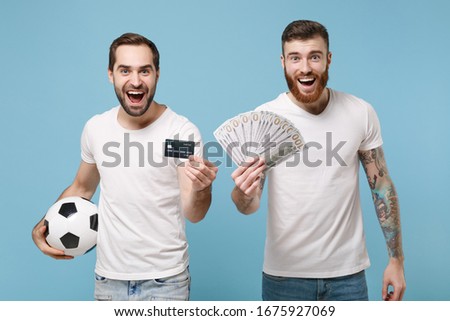 Excited two men guys friends in white t-shirt isolated on blue background. Sport leisure lifestyle concept. Cheer up support favorite team with soccer ball hold fan of cash money, credit bank card
