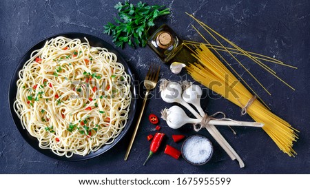 Pasta Aglio, Olio e Peperoncino, italian spaghetti with garlic, chili pepper and olive oil on a black plate with golden fork and ingredients on a concrete table, flat lay, horizontal view from above Royalty-Free Stock Photo #1675955599
