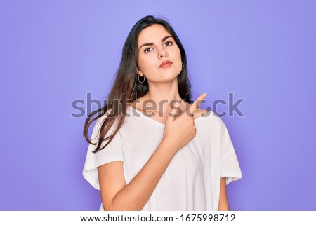 Young beautiful brunette woman wearing casual white t-shirt over purple background Pointing with hand finger to the side showing advertisement, serious and calm face