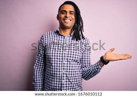 Young handsome african american afro man with dreadlocks wearing casual shirt smiling cheerful presenting and pointing with palm of hand looking at the camera.