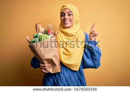 Young beautiful brunette arab woman wearing islamic hijab holding paper bag with food surprised with an idea or question pointing finger with happy face, number one