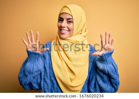 Young beautiful brunette muslim woman wearing arab hijab over isolated yellow background showing and pointing up with fingers number ten while smiling confident and happy.