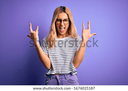 Young beautiful blonde woman wearing striped t-shirt and glasses over purple background shouting with crazy expression doing rock symbol with hands up. Music star. Heavy music concept.