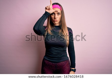 Young blonde fitness woman wearing sport workout clothes over isolated background making fun of people with fingers on forehead doing loser gesture mocking and insulting.