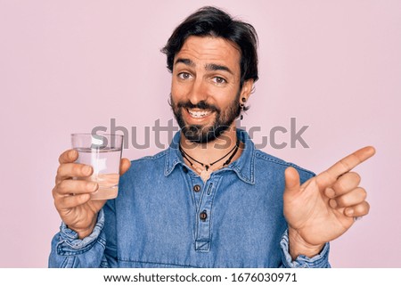 Young handsome hispanic bohemian hipster man with bear drinking a fresh glass of water very happy pointing with hand and finger to the side