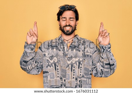 Young handsome hispanic bohemian man wearing hippie style and sunglasses gesturing finger crossed smiling with hope and eyes closed. Luck and superstitious concept.