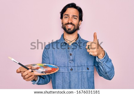 Young handsome hispanic artist man with bear holding painter palette and paintbrush happy with big smile doing ok sign, thumb up with fingers, excellent sign