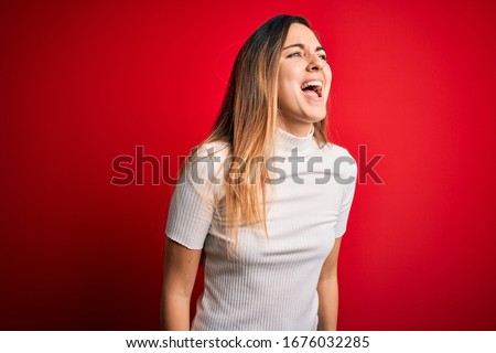 Beautiful blonde woman with blue eyes wearing casual white t-shirt over red background angry and mad screaming frustrated and furious, shouting with anger. Rage and aggressive concept.