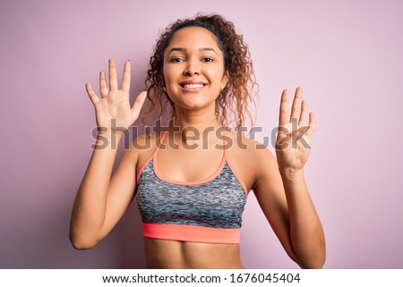 Beautiful sportswoman with curly hair doing sport wearing sportswear over pink background showing and pointing up with fingers number nine while smiling confident and happy.
