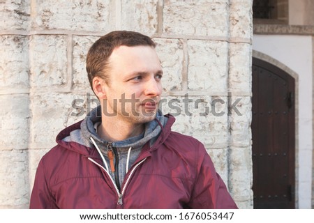 Young Caucasian man in a red sporty jacket standing near old stone wall in evening sunlight Royalty-Free Stock Photo #1676053447
