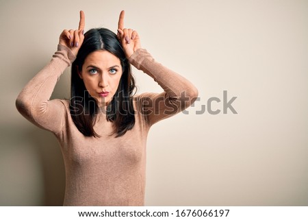 Young brunette woman with blue eyes wearing casual sweater over isolated white background doing funny gesture with finger over head as bull horns